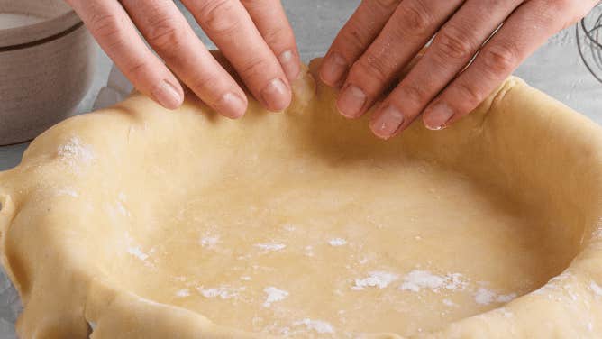 Fingers from 2 hands are touching the edge of a round dish covered with raw pastry.