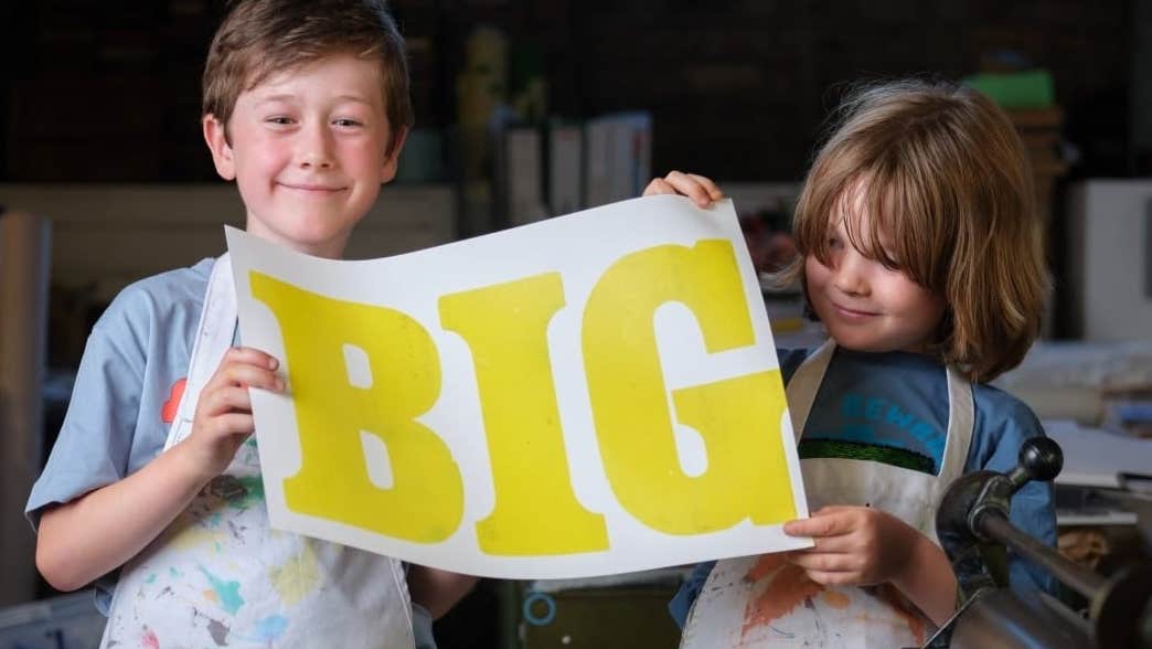 2 smiling young children holding up a large printed piece of paper with single large word on it.