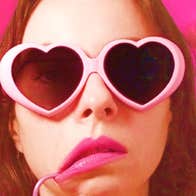 Close up head shot of woman wearing pink heart shaped sunglasses, pink lipstick and a pink straw pulling on the corner of right side of her mouth, against pink background.