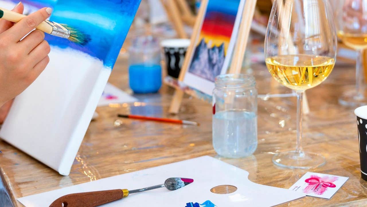 A person painting a canvas at a table with brushes and a glass of wine