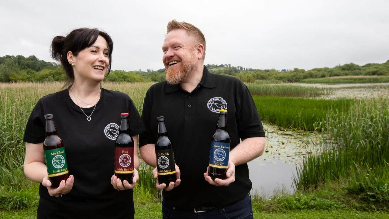 Two people showcasing a selection of beers against the backdrop of a lakeside wetland