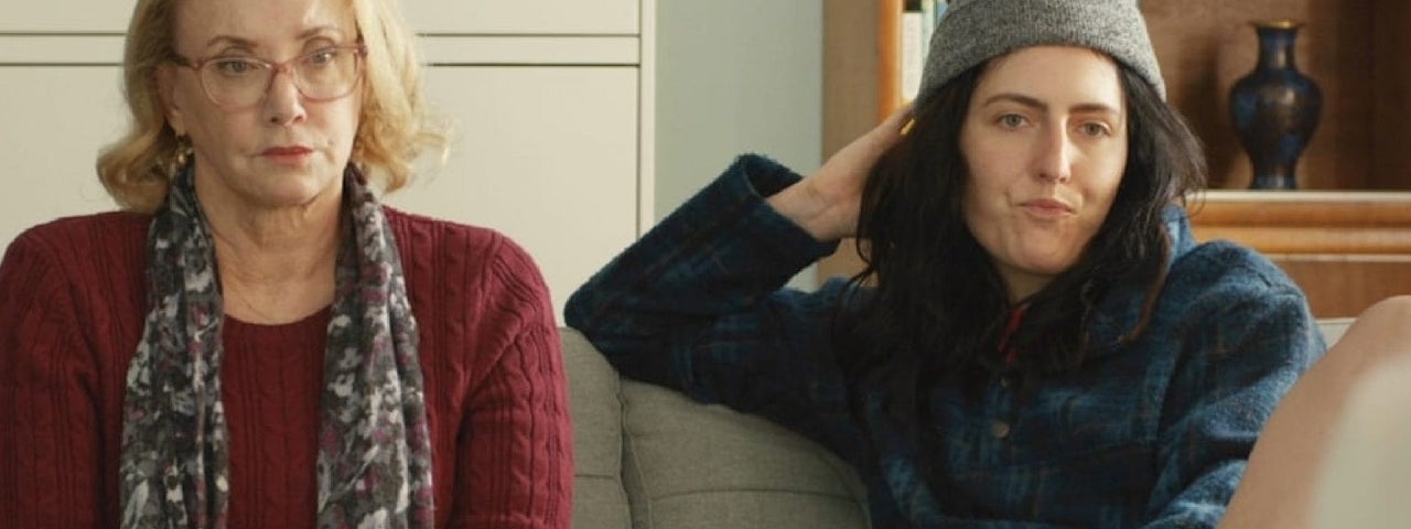 The Year Between Movie Still. 2 people are sat on a sofa, looking unhappy or sceptical.