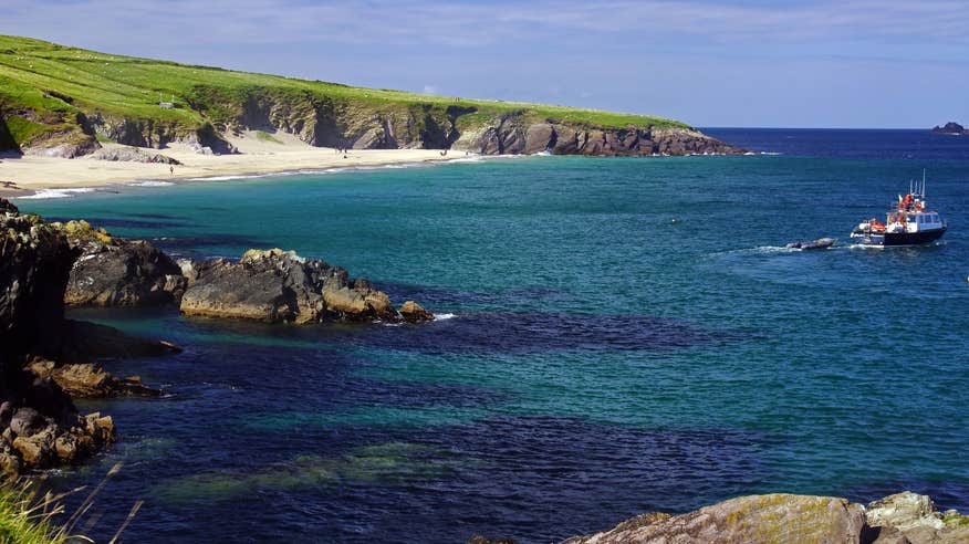 A boat gliding by a golden beach and rocky coves in the distance