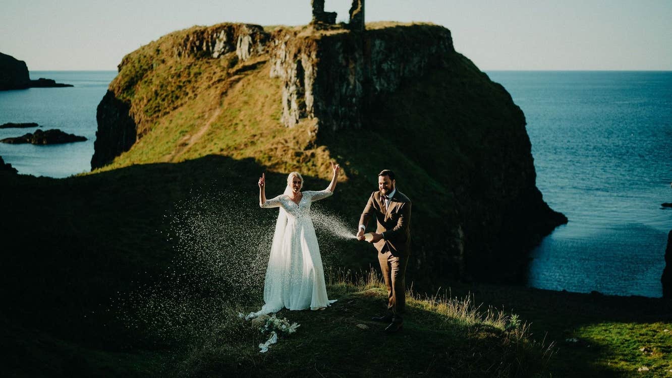 A bride and groom stand near a rocky coastline with ruins in the background
