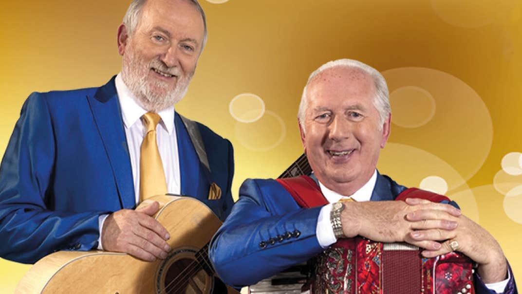 2 smiling older men resting their hands on musical instruments around their necks on straps