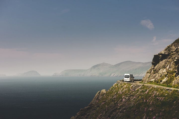 A minibus drives around a bend to the right by a cliff edge which opens out to the sea on the left