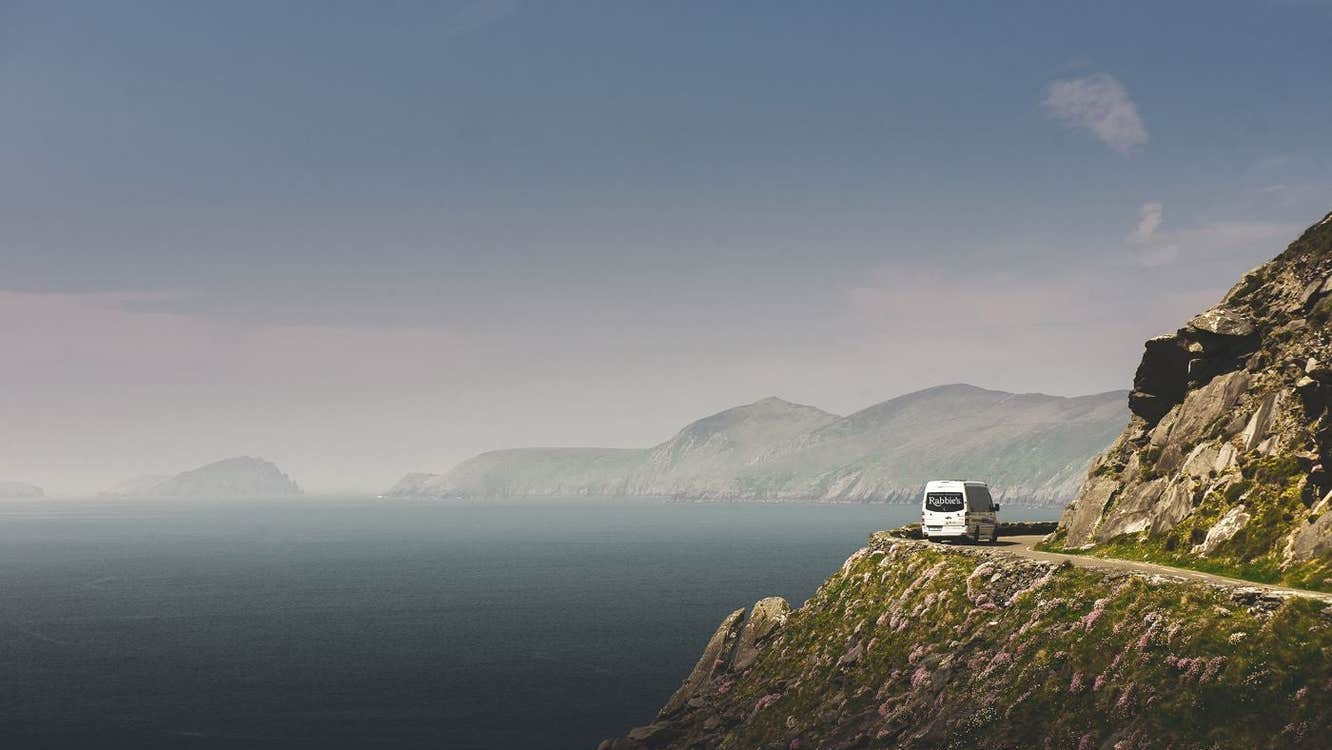 A minibus drives around a bend to the right by a cliff edge which opens out to the sea on the left