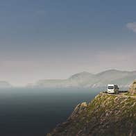 A minibus drives around a bend to the right by a cliff edge which opens out to the sea on the left