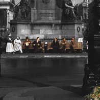 People scattered along 2 benches underneath plinth of large monument in a city in the past.