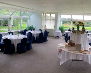 Function room with tables white tablecloths blue chairs beige carpet and windows