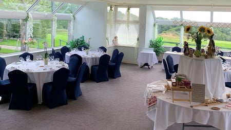 Function room with tables white tablecloths blue chairs beige carpet and windows