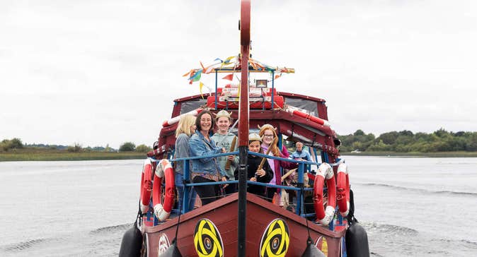 Image of Viking Ship Athlone with people standing at the bow of a small brown ship on the water.