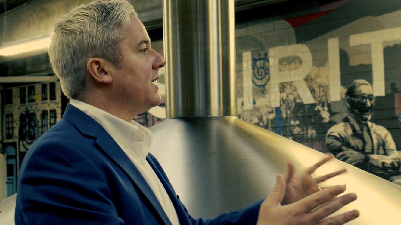 Whiskey expert standing by a nearby still addresses an unseen group of people