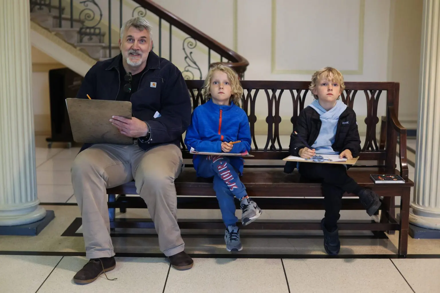 A man and 2 boys are seated on a wooden bench in a hallway with clipboards and pencils.