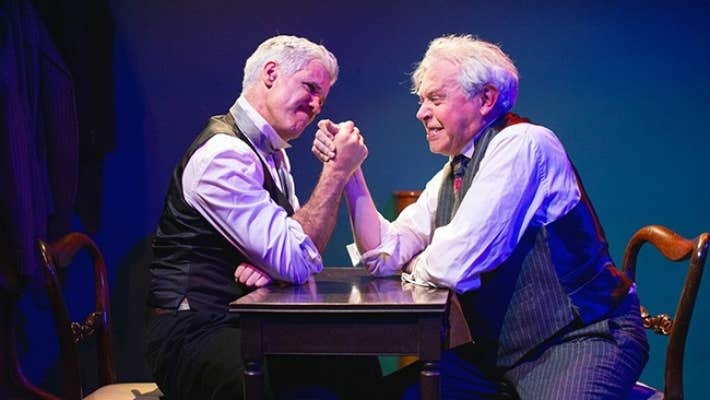 2 men in shirts and waistcoats arm wrestling on a small table.
