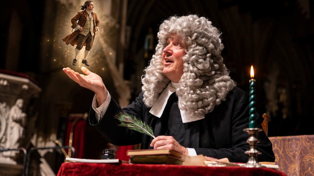 A man in white curly wig is seated at a table with a candle and a tiny figure of a man on his outstretched hand.