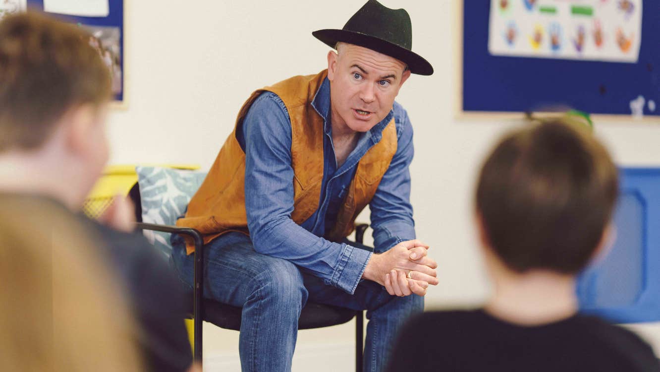 Killian Storytelling for Children - a man in a black hat, seated leaning forward talking, blurred rear view of children in foreground.