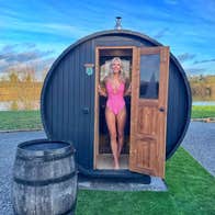 Lady in a pink swimsuit standing in the doorway of an open sauna by a lake