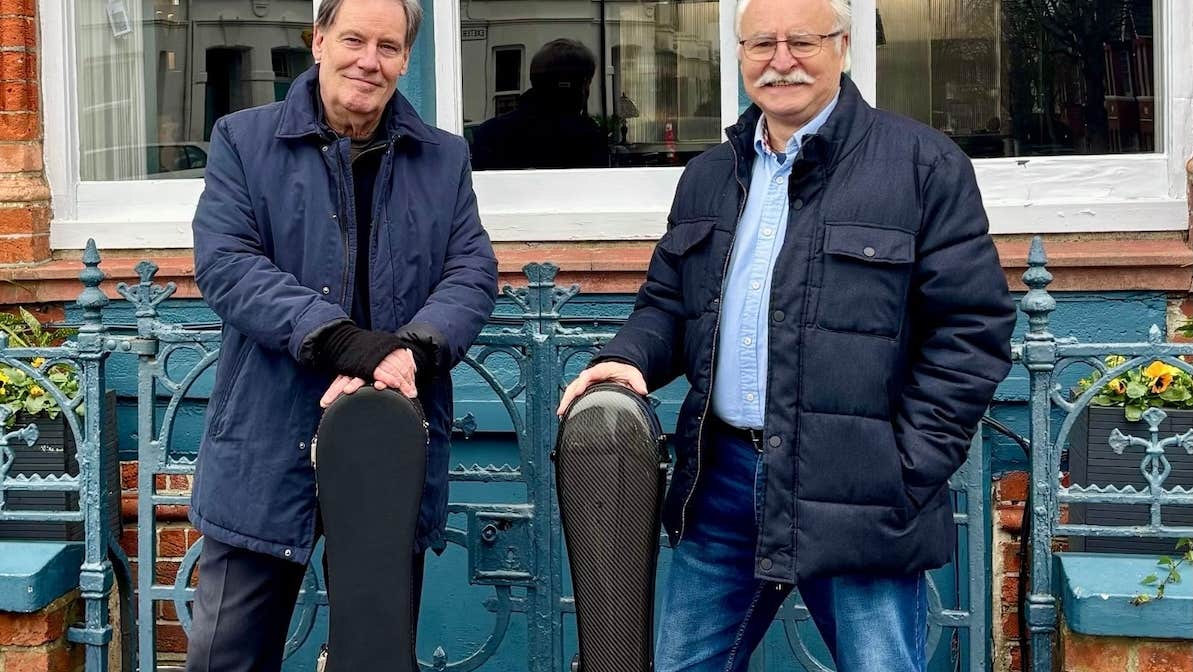 2 older smiling men with guitar cases standing in front of railings.