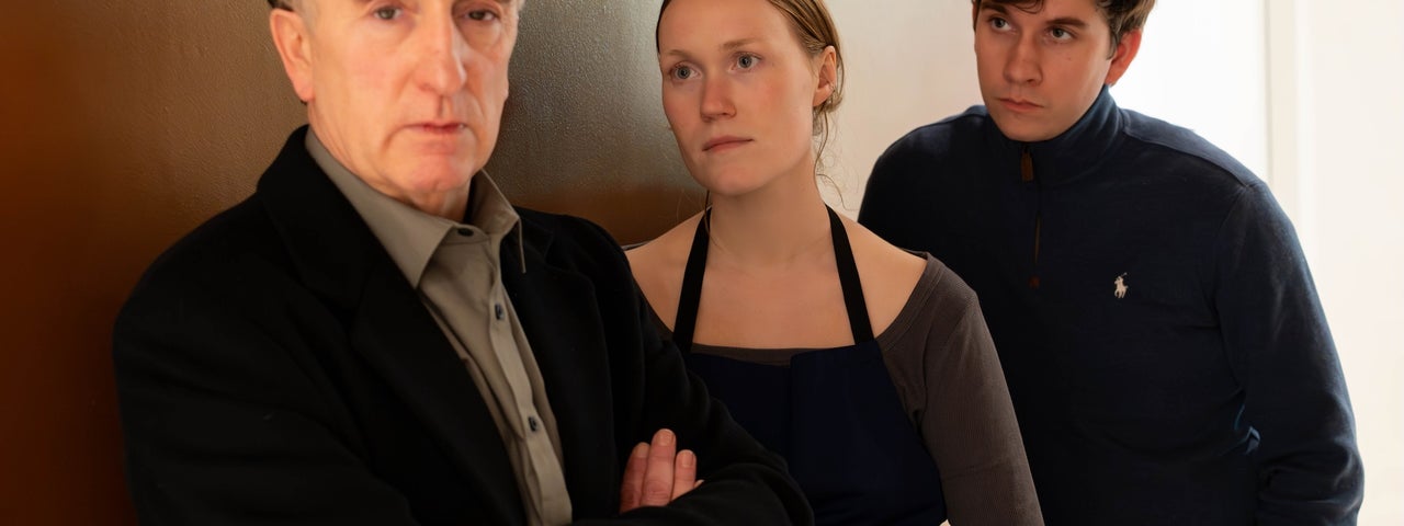 2 people are leaning against an internal wall looking at a man with his arms crossed, not looking at them.