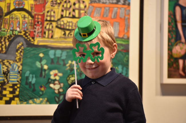 A young boy in dark top is holding up green shamrock style glasses frames on a stick in front of his face, with a nervous smile and small, green hat on his head, behind is multicoloured art work.