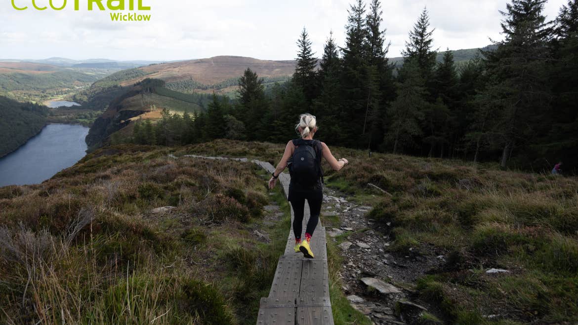 EcoTrail Wicklow 2026 - a person running along a narrow wooden path on rough land with trees and hills in background.