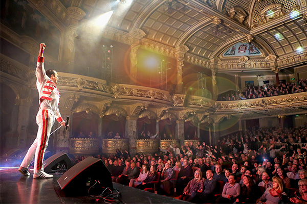 RADIO GA GA at The Everyman, a man dressed in a white with red trim outfit standing at the front of a stage with his right arm in the air, looking out over an audience in an ornate theatre.