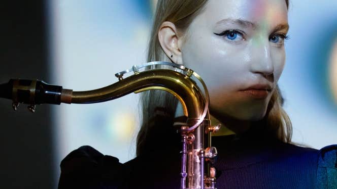 A serious looking woman in dark top with part of saxophone visible she is holding in front of her.