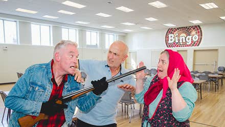 A man is holding a rifle at a horrified woman in a headscarf with another man holding him back in a hall with table and chairs.