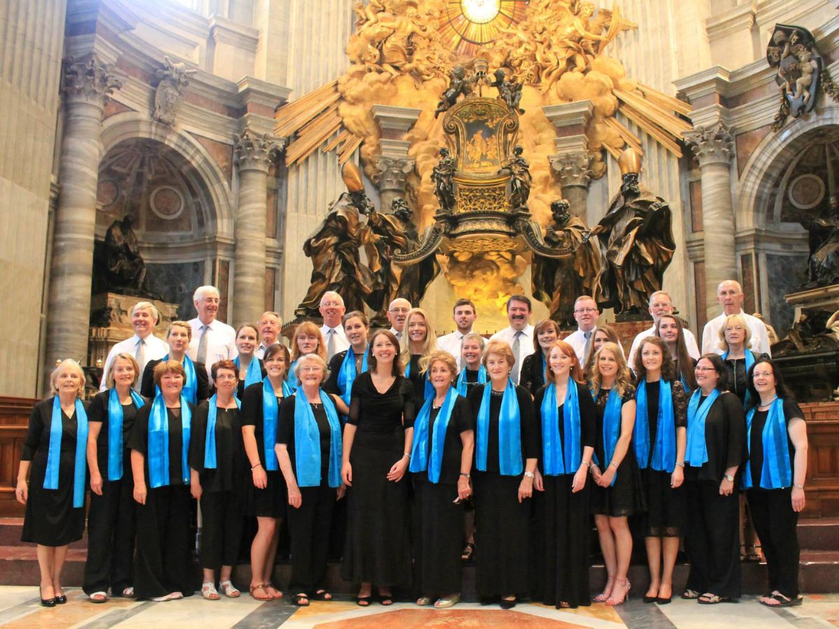 Choir group in front of an ornate gold structure in a church, the women are wearing black with light blue scarf around their necks, men in white shirts and pale ties