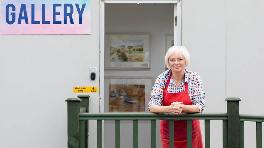 A lady leaning on a wooden fence with an open door behind her where there are pictures on a wall