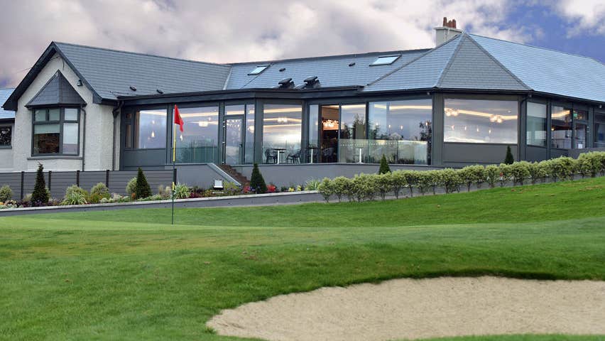 External view of the Clubhouse showing a low rise glass fronted building and overlooking bunkers on the green with a flagstick also visible on the green