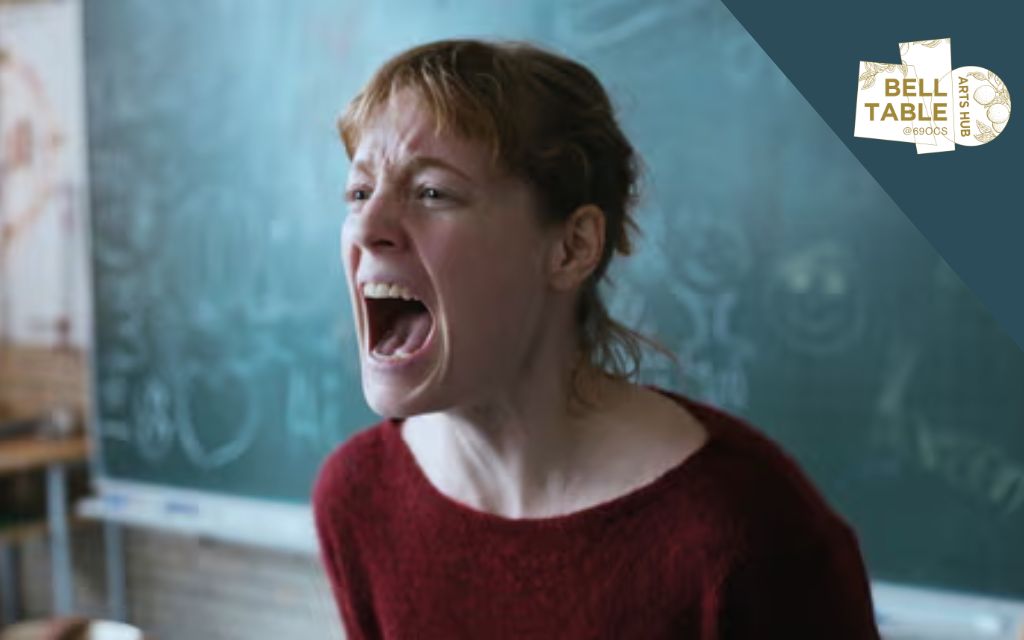 A woman is looking distressed, shouting with force, blurred blackboard behind her.