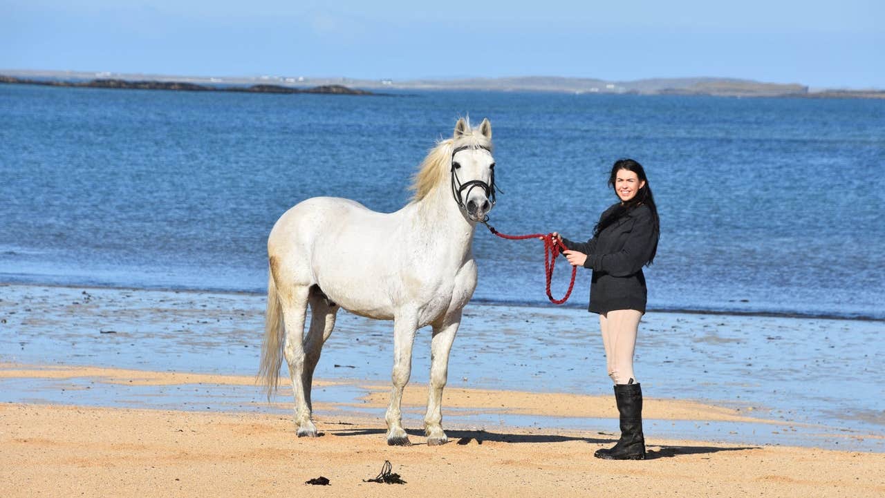 A woman in riding gear standing on a beach with a horse