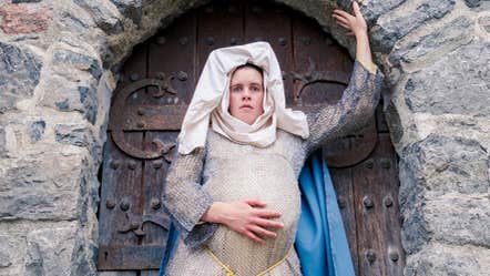 A heavily pregnant woman in medieval dress is standing in a stone archway.