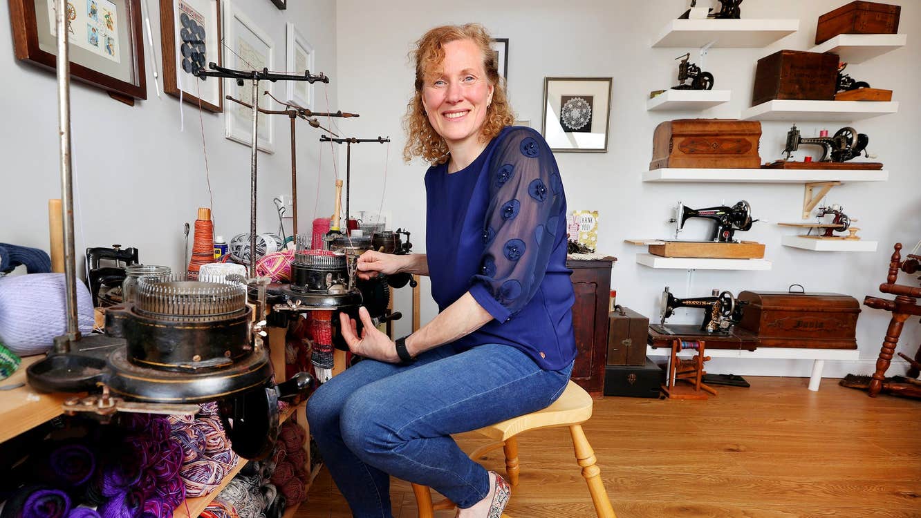 A woman sits by a knitting machine with wool under a bench