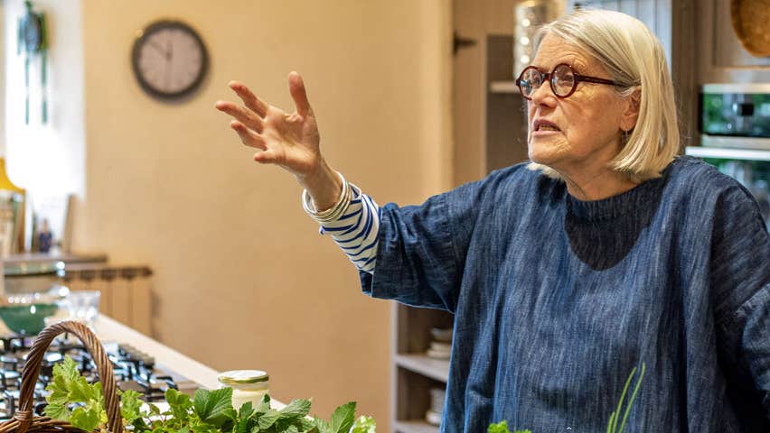 A view of Darina Allen with her right hand raised giving a talk to unseen people as she stands in front of kitchen counter with fresh herbs on display as well as a gas hob on the left hand side of the picture