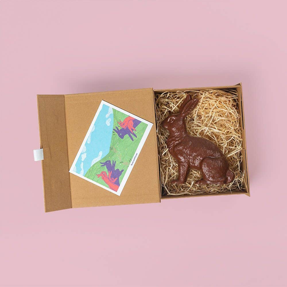 A chocolate rabbit in a cardboard box on a bright pink background, made by Bean and Goose