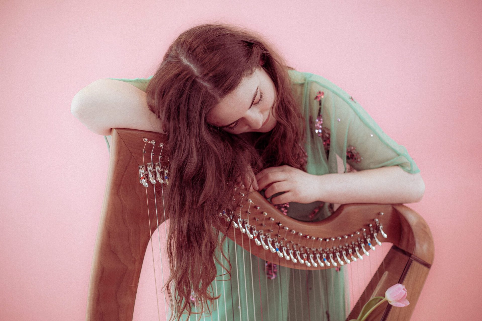 A woman in a pale green dress is leaning on the top of a harp with her hair draping over, head down looking at a flower, against pale pink background.