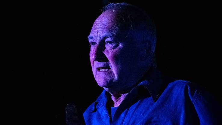 Actor Donal O'Kelly standing against a black background, face is lit by a purple-blue light