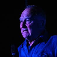 Actor Donal O'Kelly standing against a black background, face is lit by a purple-blue light
