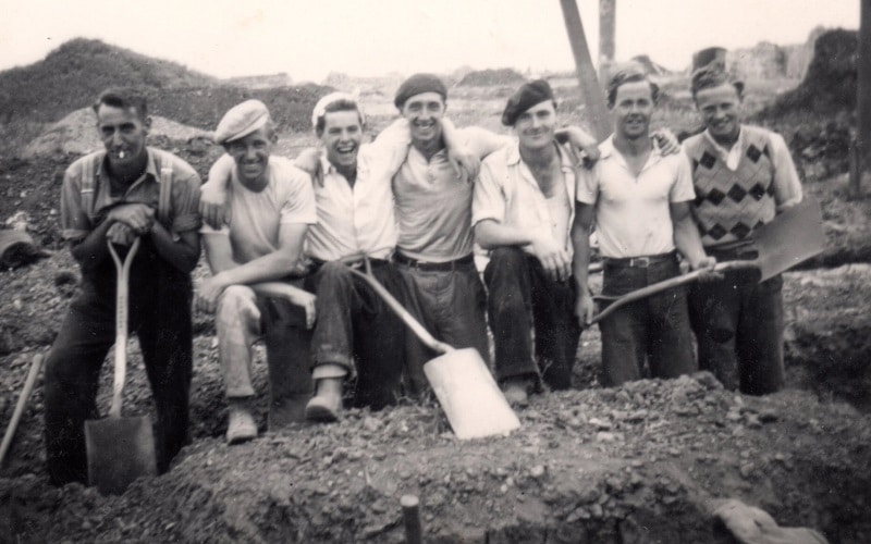 Irish Workers Pictured Together in Britain. Old black and white photo of line of 7 men in working clothes standing in a ditch, some with shovels.