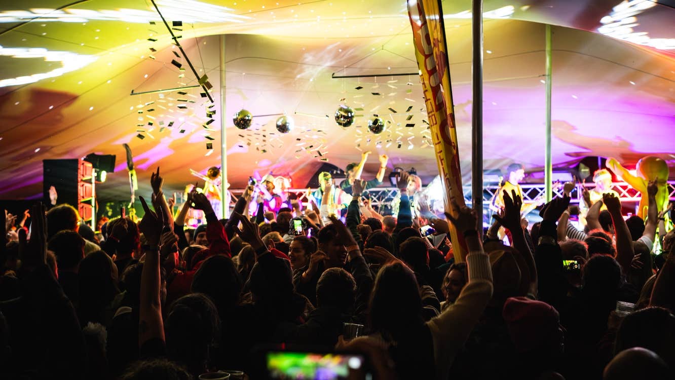 Inside a large event tent with people performing at the front, an audience in shadow and the roof lit in yellow and purple lights.