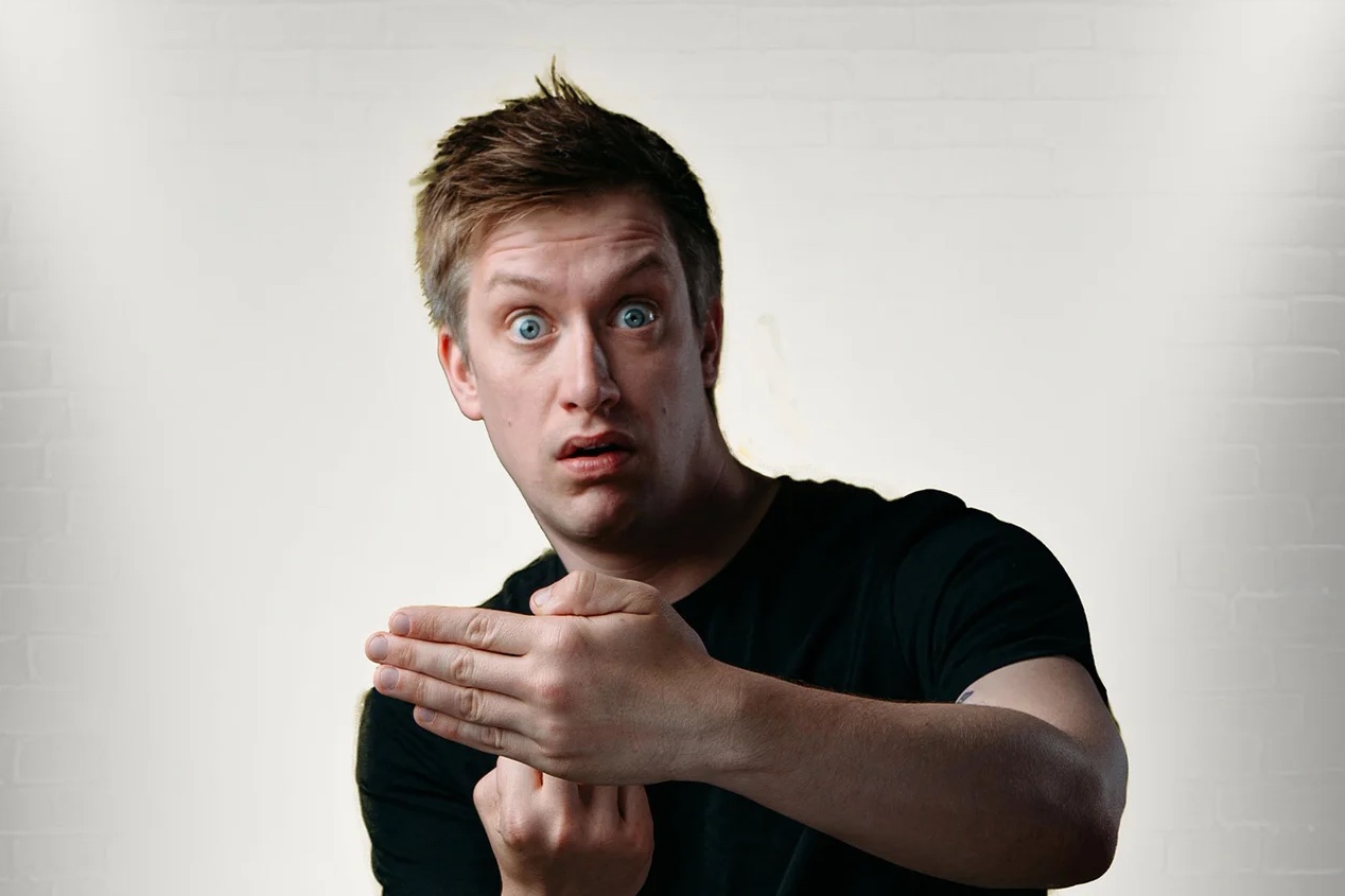A man in black T-shirt is looking shocked at camera, with implied gesture with one hand, being covered over by the other
