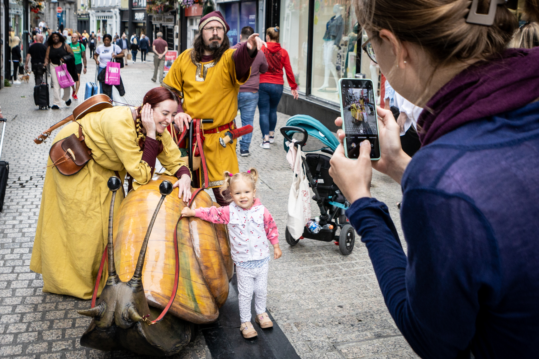 In a paved town street, a small child is having her picture taken with 2 people in mustard coloured costumes with a giant yellow snail.