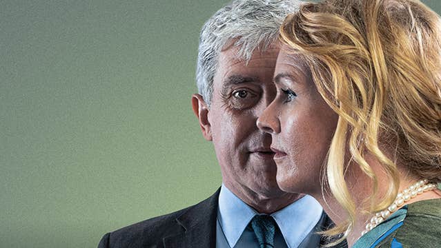 A man in a suit looking ahead with a woman in side profile slightly in front of him.