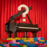 A grand piano is pictured with piles of different coloured lego bricks in the foreground and red, stage curtains behind in the background.