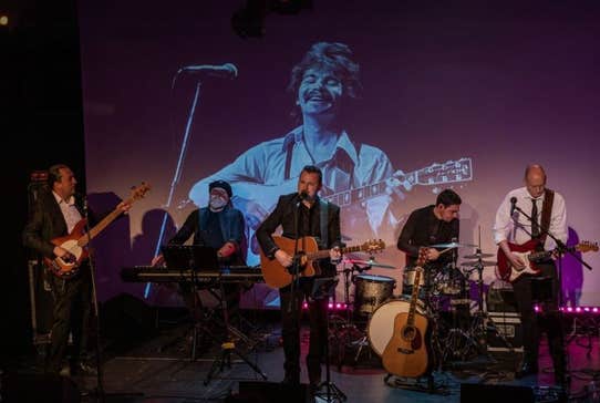 A band of 5 men playing instruments on a small stage with backdrop of image of a man playing a guitar