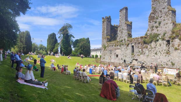 Free Open Air Recital Castlemartyr, people sat in chairs spread across a sloping green area looking at group performing against background of ruined castle.