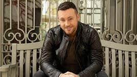 A smiling man dressed in black seated on a bench in front of metal railings